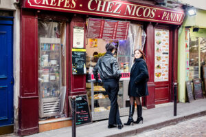 Christian and Alicia stand in front of Chez Nico's, a tempting food stand offering crepes and panini. Christian sports a black leather jacket, while Alicia opts for a long black coat and boots. The inviting menu showcases an array of delicious options.