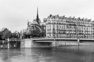 Black and white image of the Seine River in Paris with the Notre-Dame Cathedral visible in the background. A bridge crosses the river, and classic Parisian buildings are seen along the riverside. Cloudy sky adds a moody atmosphere.