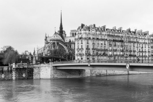 Black and white image of the Seine River in Paris with the Notre-Dame Cathedral visible in the background. A bridge crosses the river, and classic Parisian buildings are seen along the riverside. Cloudy sky adds a moody atmosphere.