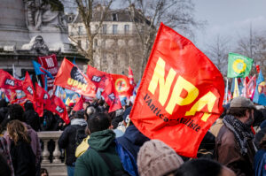 A crowd of people at an outdoor protest in Paris, holding various colorful flags and signs, including a prominent red flag with "NPA" in yellow letters. The Place de la République is bustling under a cloudy sky, embodying the spirit of a Manifestation Antifa. Trees and buildings frame the scene.