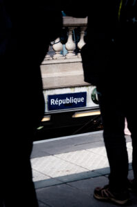 View of a "République" metro sign through the legs of two people on a platform, partially framed by a balustrade. Amid echoes of "No Pasaran," the scene captures the atmosphere at Place de la République, where voices gather for manifestation Antifa.