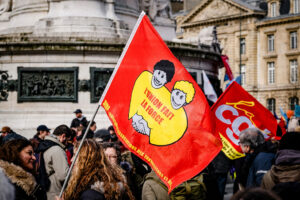 Protesters gather, holding colorful flags in a lively Manifestation Antifa. One red flag features two people embracing with the phrase "L'union fait la force," reminding all that "Unity is strength." This powerful scene unfolds against the backdrop of a historical building in Paris.