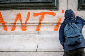 A person in a hooded jacket and backpack spray-paints orange graffiti letters on a stone wall in Paris. The partially visible word begins with "ANTI," hinting at the spirit of a manifestation.