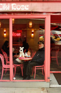 A man sitting at a table in a café with red chairs and decor, looking out through the window. Next to him are figures of a white dog and a boy from a comic series with a sign reading "Milou." Neon lights illuminate the interior.