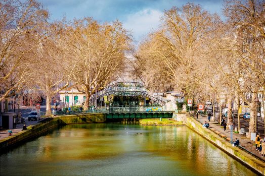 A scenic canal lined with bare trees and bordered by walkways. People sit on benches along the water. In the background, a green cast-iron footbridge arches over the canal under a partly cloudy sky. Ducks swim in the water.