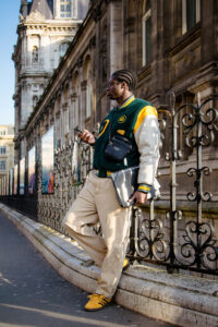 Valentin Robert, in his green and yellow varsity jacket, leans nonchalantly against a fence, juggling a phone and laptop. He sports beige pants and yellow shoes, his braided hair complementing the backdrop of a historic stone building and a row of photo displays.