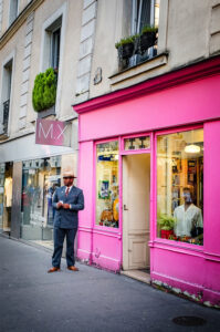 A man in a suit and hat, reminiscent of Cedric Bidault's distinct style, stands outside a bright pink storefront on the bustling city sidewalk. Mannequins pose in the window beside him, surrounded by neighboring shops and a light-colored building adorned with lush plant decorations above.
