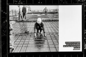 Black and white image of construction workers laying rebar for the Tour Montparnasse in Paris, 1971. A worker in the foreground kneels, handling rebar, while others toil in the background. Captured with Gérald Bloncourt's eye for detail, a sign identifies the workers as Portuguese.