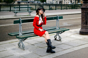 A woman in a robe rouge, black boots, and a chapeau noir sits on a green bench near a street. She is smiling, wearing sunglasses, and appears relaxed in this urban outdoor setting.