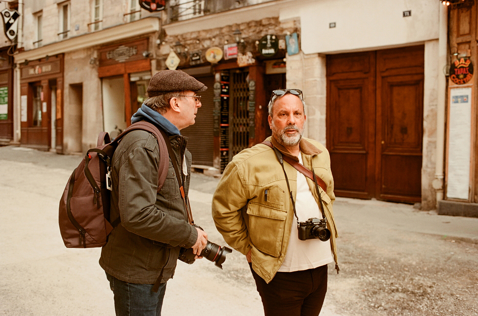Two men stand on a street, both holding cameras. One wears a cap and glasses, the other wears a light jacket. Behind them are storefronts with closed wooden doors. The street appears quiet and empty.