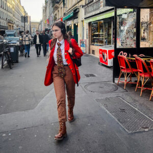 A person strolls down a city street, donning a vibrant red coat reminiscent of Angeline Girardot's iconic style, paired with brown pants and boots. Headphones in place, they pass outdoor cafes and bustling shops, while other pedestrians and parked bicycles animate the backdrop.