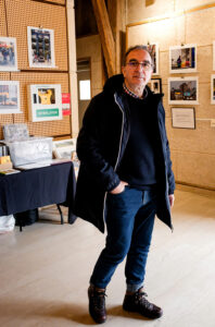 A man in a black jacket, sweater, and jeans stands at the 2025 Expo Photos Onzain exhibition. Photo displays adorn the gallery walls, while a table labeled "Defibrillator" sits in the background with various items.