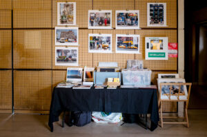A table draped in a black cloth showcases assorted books and photographs, embodying the spirit of Expo Photos Onzain. The wall behind features a collection of framed photos capturing urban scenes, while a green "Defibrillator" sign is visible on the right.