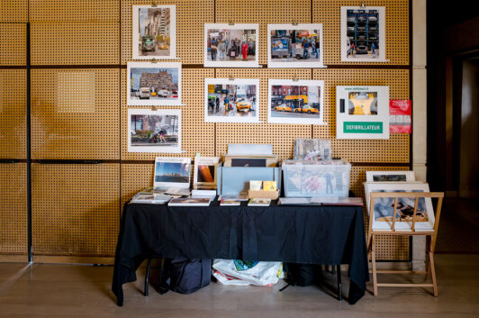 A table draped in a black cloth showcases assorted books and photographs, embodying the spirit of Expo Photos Onzain. The wall behind features a collection of framed photos capturing urban scenes, while a green "Defibrillator" sign is visible on the right.