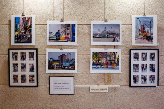 A display of framed photos arranged on a textured wall showcases urban scenes, from street performers to autumn cityscapes. A small sign reads "New York, New York," and a name tag is visible below, part of the captivating Expo Photos Onzain running from 15/03/2025 to 23/03/2025.