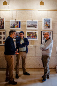 Three men stand talking in front of a wall displaying framed photographs at the 2025 Expo Photos Onzain. They are casually dressed, and the setting exudes the vibrant atmosphere of an art exhibition or gallery.