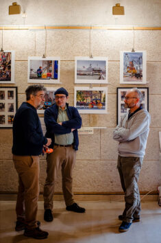 20250315-DSCF1857-Enhanced-NR_MOD_FRAME_resize Three men stand talking in front of a wall displaying framed photographs at the 2025 Expo Photos Onzain. They are casually dressed, and the setting exudes the vibrant atmosphere of an art exhibition or gallery.