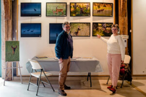 Two people stand smiling in a rustic gallery at Expo Photos Onzain. They point at nature photographs displayed on the wall featuring deer in various settings. A table with more prints is in front of them, and a small easel holds an additional photo.