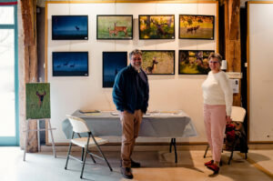 Two people stand in front of a stunning Expo Photos Onzain display, showcasing wildlife photos featuring deer in natural settings. A small table with brochures and an easel with another captivating photo are in the foreground, set within the gallery's wooden-beamed space.