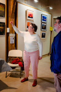 A woman in a white sweater and pink pants gestures towards the photos at Expo Photos Onzain, discussing them with a man in a blue jacket. The gallery walls showcase framed photographs under warm lighting. Behind them rests a chair holding several items.