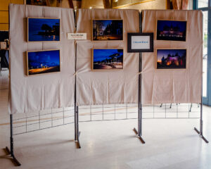 A display stand with beige fabric holds six framed landscape photographs, capturing scenic views mostly during twilight or nighttime. Featured in the Expo Photos Onzain on 23/03/2025, the setting is indoors, illuminated by natural light from a nearby window.