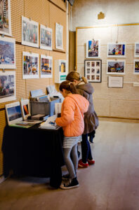 Two people stand in a gallery during the Expo Photos Onzain, viewing photographs displayed on walls. One of them, in an orange jacket, examines a book on a table with more materials. The room's light wooden flooring and pegboard-textured walls add to the event's charm.