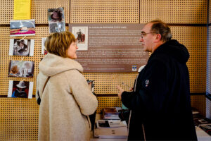 Two people engage in conversation in front of a display board with historical content at the March 2025 Onzain event. The background features a wooden pegboard with Expo photos of hats and other objects, while books and pamphlets rest on a table below the display.
