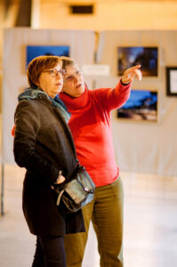 Two people stand in an art gallery at the Onzain photo exhibition. One wears a red sweater, pointing towards a piece of artwork while the other listens attentively. Blurred framed photos and a white backdrop hint at the much-anticipated 2025 exhibition dates.