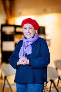 An older adult stands indoors, smiling warmly at the Onzain event. They wear a red hat, a purple scarf, and a dark sweater. The background is softly blurred, showing chairs and a bulletin board from the Expo Photos Onzain. The atmosphere is cozy and welcoming.