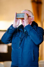 An older person stands indoors at the Expo Photos Onzain 2025, wearing a blue winter jacket and holding a rectangular device to their eyes, seemingly immersed in the exhibition. The softly blurred background adds an air of mystery.