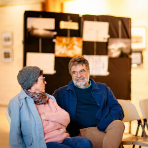 An older man and woman sit together in conversation at Expo Photos 2025. The man wears glasses and a blue jacket, while the woman dons a gray hat and pink sweater. Behind them, blurred images reminiscent of Onzain are displayed on a black stand.