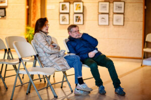 A man and woman sit in plastic chairs, casually dressed in jackets. The man leans back with his legs crossed, while the woman sits upright. They are surrounded by framed photos on the wall, perhaps from the Expo Photos Onzain, with other empty chairs around them.