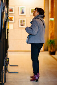At the Expo Photos Onzain 2025, a woman in a gray coat and purple shoes admires framed pictures in the art gallery. The beige walls behind her are adorned with diverse artwork. She carries a light-colored bag and stands gracefully on the polished floor.