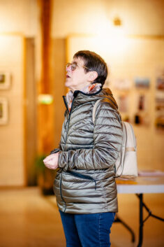 A person in a metallic puffer jacket, blue jeans, and a beige backpack stands indoors at the warmly lit Expo Photos Onzain. Blurred photos or artwork adorn the walls as they look slightly upward, immersed in thought.