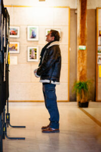A person in a leather jacket and blue pants stands in an art gallery, captivated by framed pictures at the Onzain Expo. A leafy plant accents the beige walls and wooden beam backdrop, enhancing the serene atmosphere of this remarkable photo exhibition.