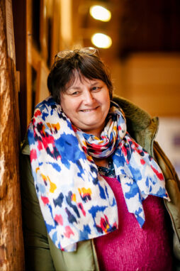 A person with short, dark hair is wearing glasses on their head, a colorful patterned scarf, and a green jacket. The warm indoor lighting adds to the charm as they smile, leaning against a wooden surface at the Expo Photos Onzain.