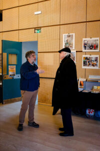 Two men stand in conversation in a wood-paneled room, part of the Expo Photos Onzain exhibition. One gestures animatedly while the other, clad in a black coat and hat, listens intently. Posters adorn the walls, and a table with books stands nearby. To the left, an open blue door beckons toward 2025’s innovative displays.