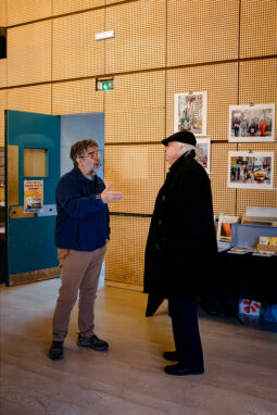 Two men stand in conversation in a wood-paneled room, part of the Expo Photos Onzain exhibition. One gestures animatedly while the other, clad in a black coat and hat, listens intently. Posters adorn the walls, and a table with books stands nearby. To the left, an open blue door beckons toward 2025’s innovative displays.