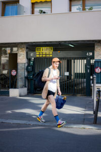 Rémi Villette, wearing a cap, beige shirt, white T-shirt, and shorts, walks on a city sidewalk carrying a bag and crate while looking at their phone. An apartment building and a sign are visible in the background.