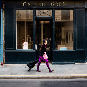 A person in a pink skirt and purple tights walks past the storefront of Galerie Grès, where artwork by Isabelle David is displayed in the large windows, pulling a wheeled bag along the city sidewalk.