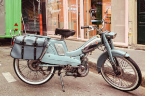 A vintage blue moped with saddlebags is parked on a city street, angled toward the sidewalk. Storefronts and a green truck are visible in the background.
