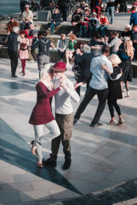 Multiple couples dance closely in an outdoor square, with onlookers seated on steps in the background. The foreground features a woman in a burgundy dress dancing with a man in a striped shirt and red hat.