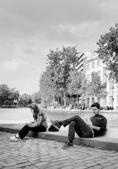 A young woman and a young man sit apart on a cobblestone edge beside a canal. The woman looks down, while the man reads a newspaper. Trees and buildings line the water in the background under a cloudy sky.