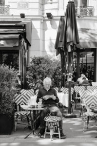 Sebastien Lemoine, an older man, sits alone at a Parisian café table reading a newspaper. Empty chairs surround him beneath a large umbrella. The outdoor café features lush plants and a "Place de la Bastille" sign in the background.