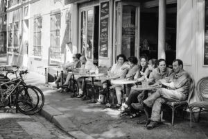 A group of people sit at outdoor tables of a café on a city sidewalk, engaged in conversation. Bicycles are parked nearby. The scene is in black and white.