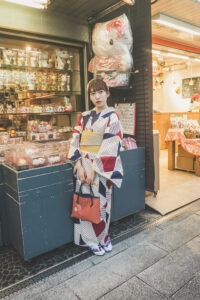 A woman wearing a patterned kimono stands on a street in front of Harika Fushima’s shop, which displays various items, including plush toys. She holds a red purse and looks at the camera, with the shop’s interior visible behind her.