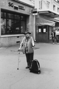 An elderly person with a cane and a shopping trolley stands on a city street in front of a Société Générale bank. Captured by Elise Vignon, the black and white photo features other pedestrians and storefronts in the background.