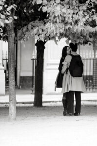 Anais and Noel stand close together under leafy trees, sharing a kiss in a park. Both wear coats and backpacks. The scene is black and white, with a building and fence visible in the background.