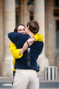 A couple embraces closely outdoors. The woman in a yellow sweater has her eyes closed, while the man in a navy sweater—Davina & Graham’s signature look—holds her with an umbrella clipped to his back pocket.