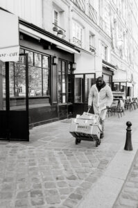 A man wearing a coat and gloves pushes a cart loaded with boxes along a cobblestone sidewalk outside a café in a city. The scene is in black and white.